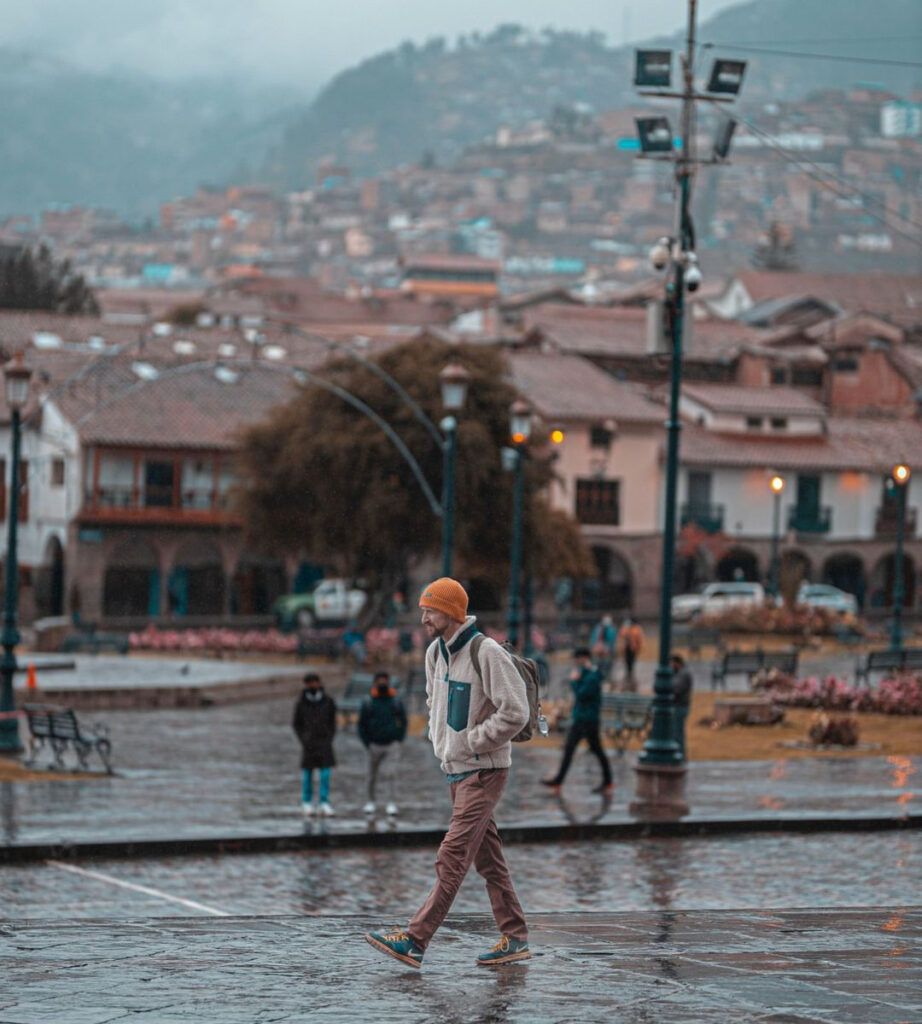 Turista caminhando pela Praça de Armas de Cusco em um dia nublado de janeiro, com casas coloniais e montanhas andinas ao fundo, experiência cultural autêntica na cidade de Cusco, Peru.