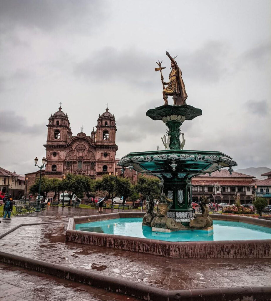 Fonte da Plaza de Armas de Cusco com estátua inca no topo, cercada por arquitetura colonial histórica, céu nublado e piso molhado após chuva, marco cultural e turístico do centro histórico de Cusco, Peru.