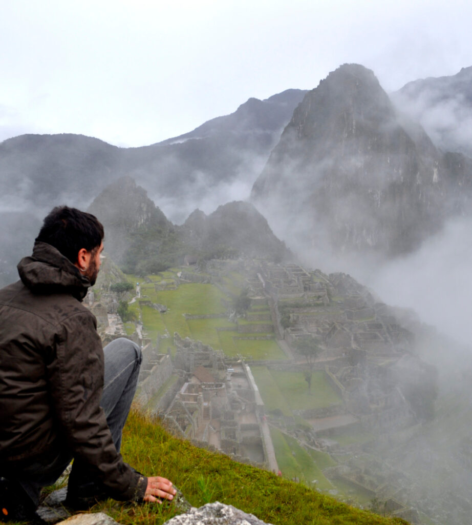 Homem observando Machu Picchu em meio à neblina durante a temporada de chuvas em janeiro, com as montanhas andinas ao fundo, experiência de viagem cultural em Cusco, Peru.