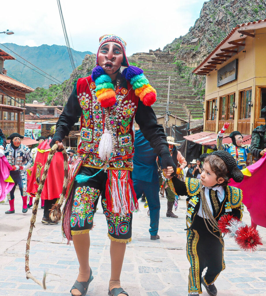Dançarino tradicional da Baixada de Reis em Ollantaytambo, no Vale Sagrado dos Incas, desfilando com trajes coloridos ao lado de uma criança durante a festa cultural de janeiro no Peru, com casas coloniais e montanhas ao fundo.