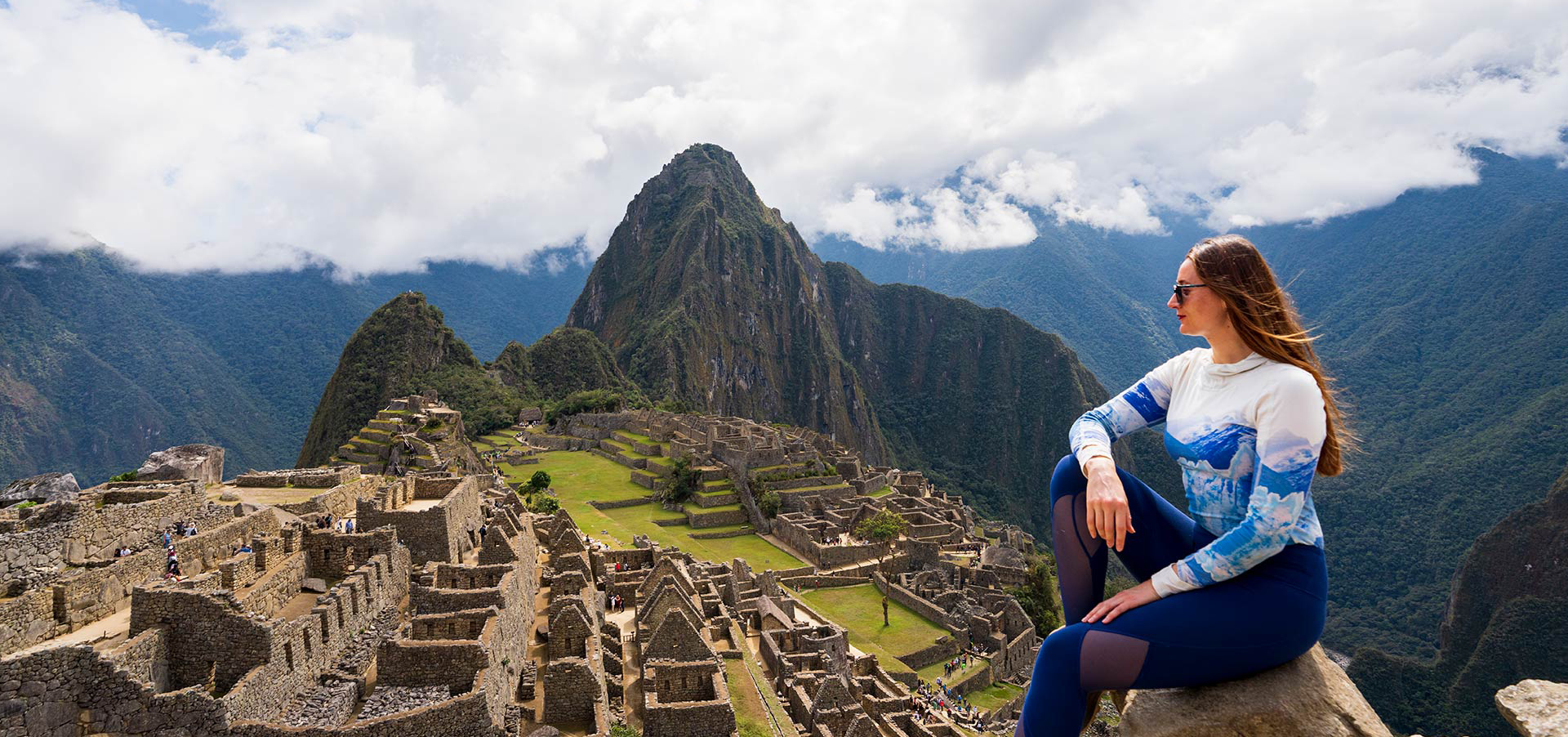 mulher sentada em uma pedra, no alto, olhando para a cidadela inca de Machu Picchu, com a famosa montanha Huayna Picchu ao fundo