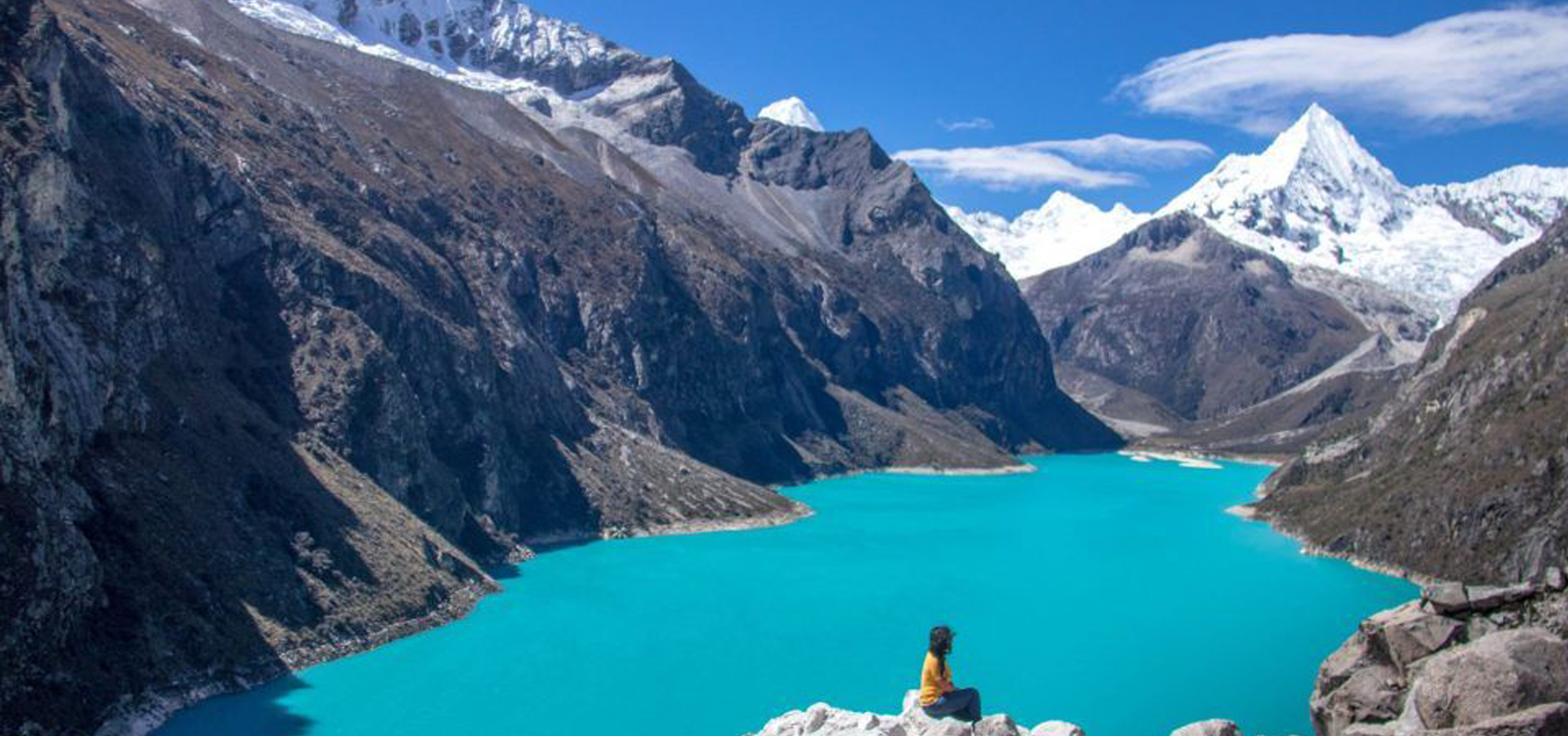 Pessoa sentada em uma rocha observando uma lagoa de águas azul-turquesa, Laguna Parón em Huaraz no Peru, cercada por montanhas altas e picos nevados sob céu azul.