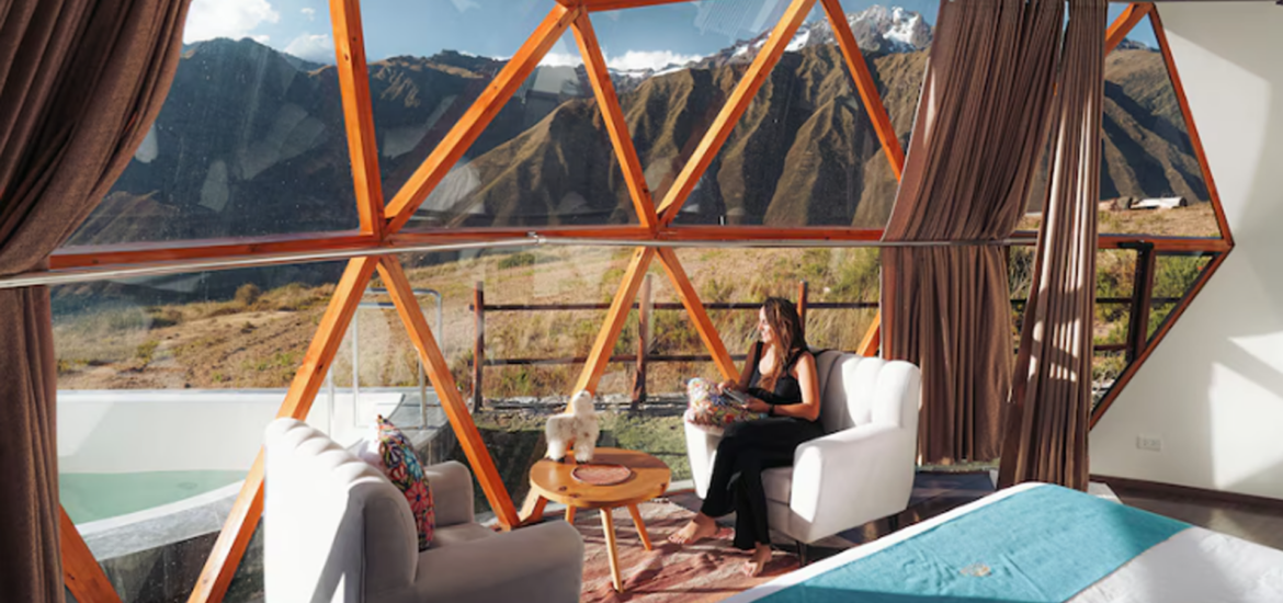 Interior de um domo de vidro com estrutura geométrica, mostrando uma mulher sentada em poltrona, com vista panorâmica para montanhas no Vale Sagrado, no Peru.