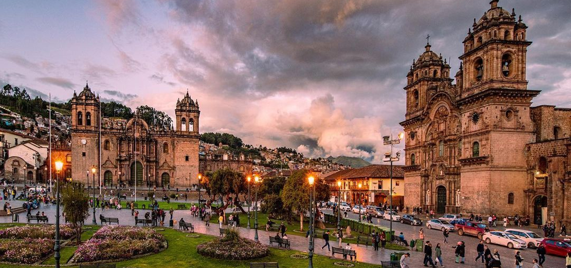 Vista panorâmica da Plaza de Armas de Cusco ao entardecer, com a Catedral de Cusco e a Iglesia de la Compañía de Jesús iluminadas enquanto moradores e turistas caminham pela praça.